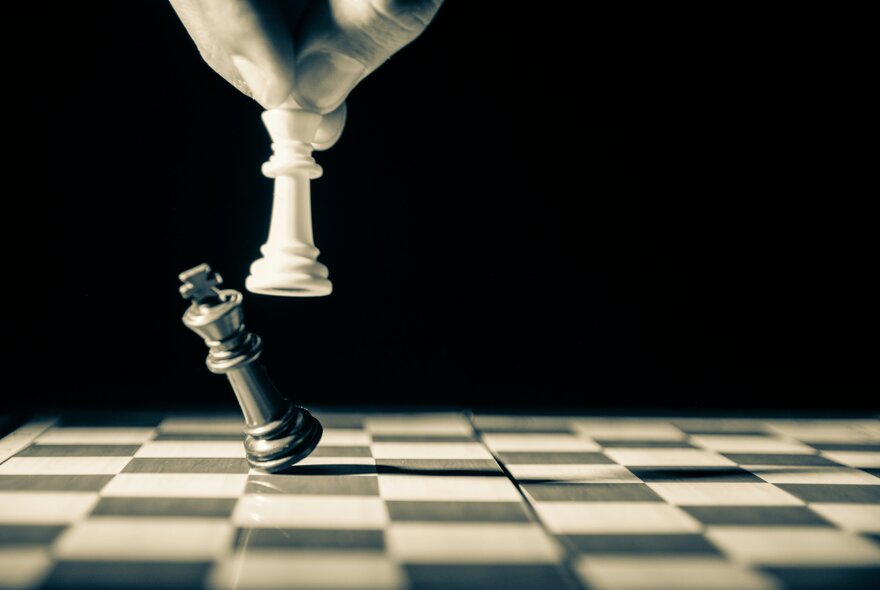 A hand holding a white chess piece, knocking over a black chess piece on a chessboard.