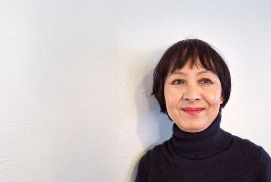 Poet Grace Yee, a smiling middle-aged woman wearing a black turtle neck jumper, with a short bowl style haircut, posed against a white background.