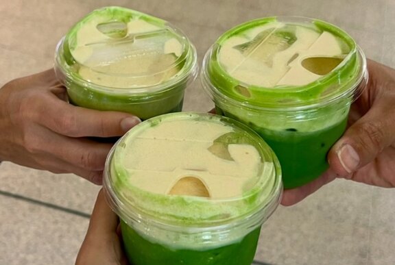Three hands each holding a matcha latte in a clear plastic cup with a clear plastic lid.