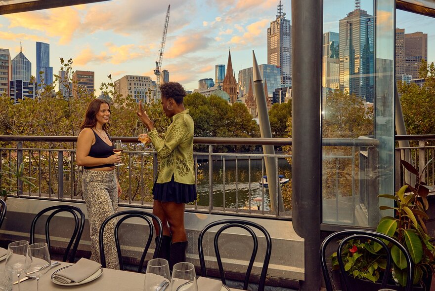 Two women standing on the balcony of a restaurant looking over the river and city skyline at sunset.