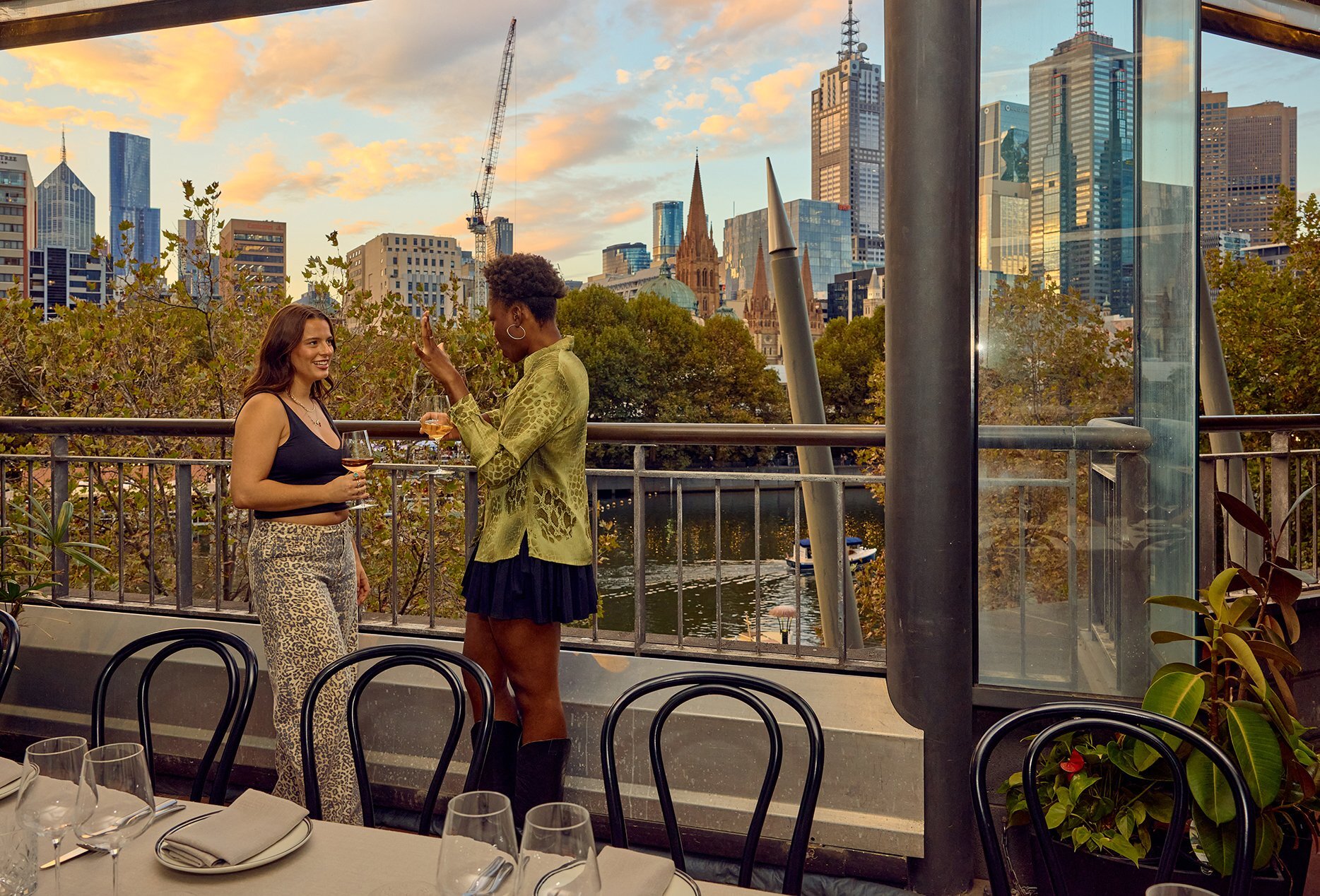 Two women standing on the balcony of a restaurant looking over the river and city skyline at sunset.
