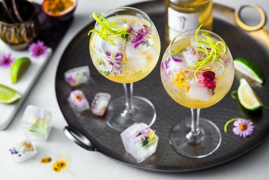 Looking downwards at a tray featuring two cocktails with flower and peel garnishes and ice cubes with flowers inside them scattered around. 