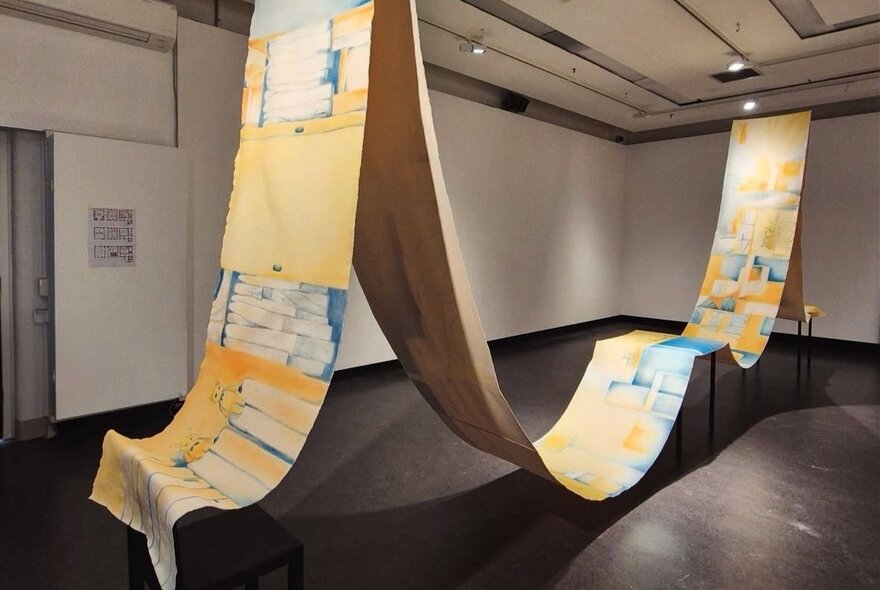 A long painting made with acrylic on starched canvas that is suspended from a gallery ceiling and two points, and balanced on two small ground supports.