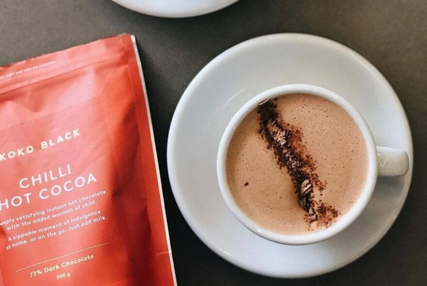 A hot chocolate on a saucer with an orange chilli hot cocoa packet next to it. 