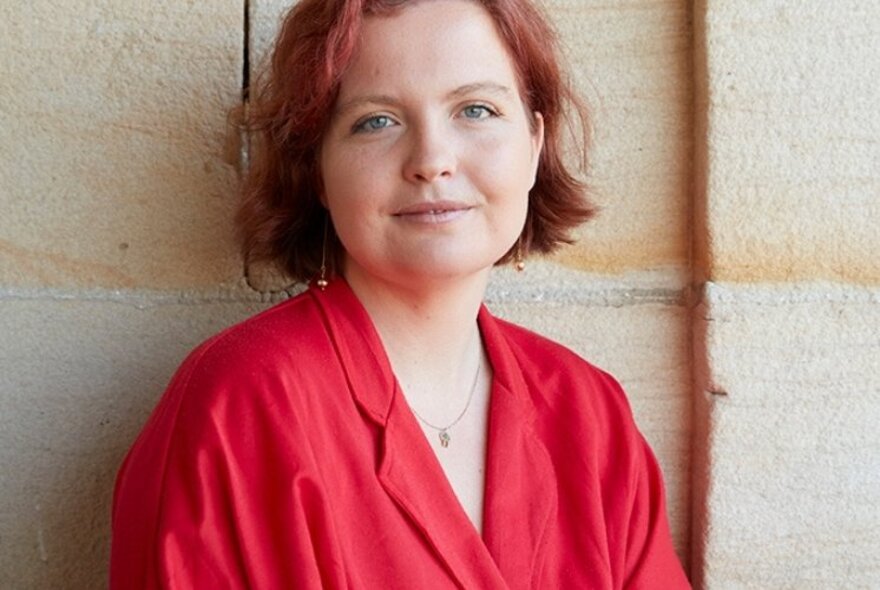 Author Madeleine Gray, a young woman with reddish hair in a bob style, wearing a red shirt and leaning against a sandstone wall. 