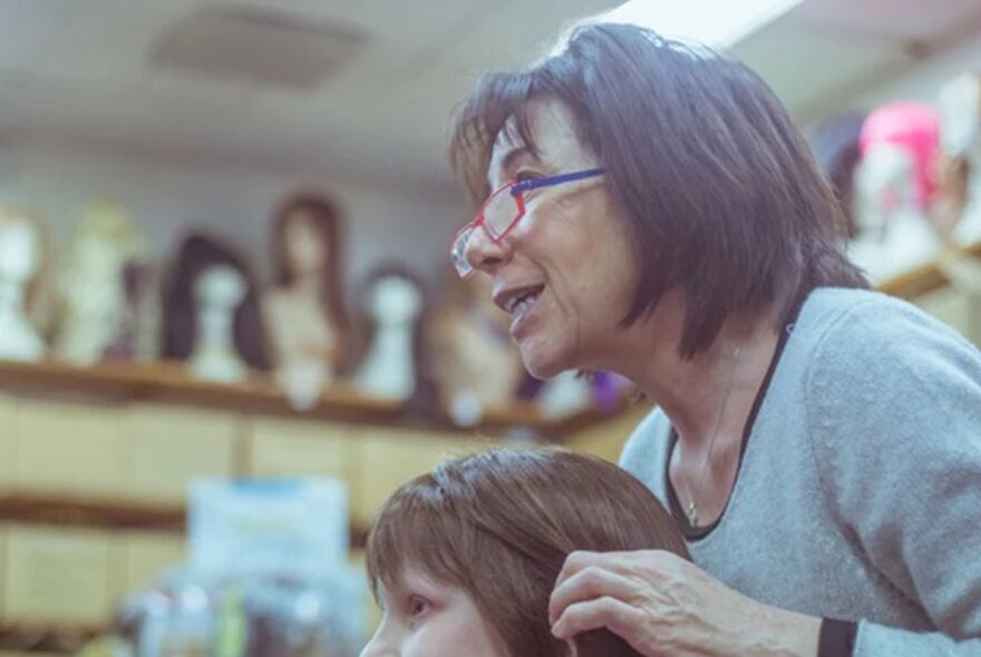 A woman fitting a wig onto another woman's head in a room with blurred wigs in the background. 