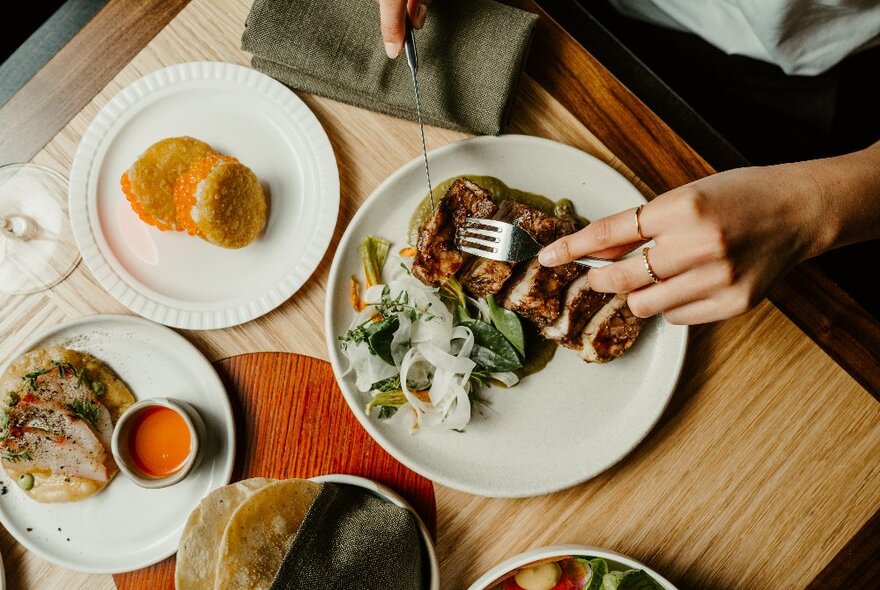 A hand using a knife and fork to cut into a steak on a plate, other dishes of food also on the dining table.