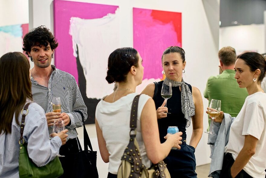 People holding glasses of wine in a bright gallery space with large abstract artworks on the walls.