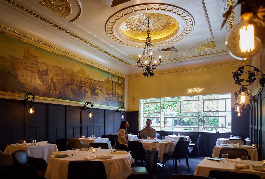 A couple dining in a sophisticated restaurant with a large mural and chandeliers.