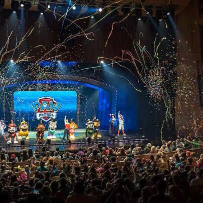 Characters from the PAW Patrol TV series performing live on a theatre stage, with streamers in the air above them and a large audience watching them.