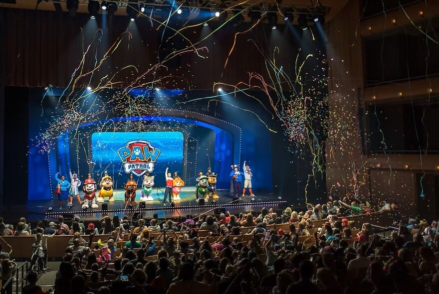 Characters from the PAW Patrol TV series performing live on a theatre stage, with streamers in the air above them and a large audience watching them.