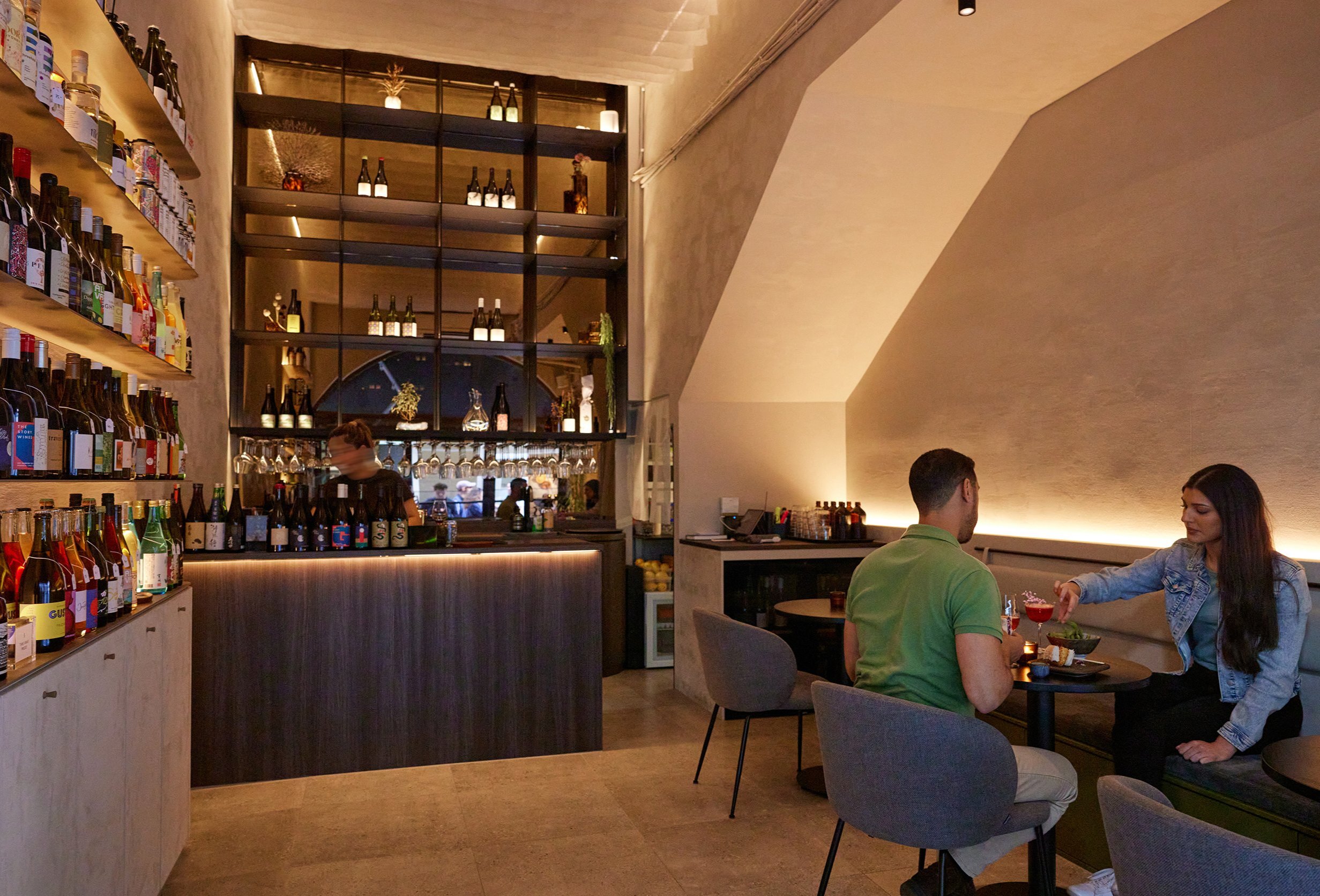 Two friends are sitting in a modern dimly lit wine bar drinking wine. There is a bar tender behind a small bar serving drinks and a shelf to the left of the bar with wine bottles on it.