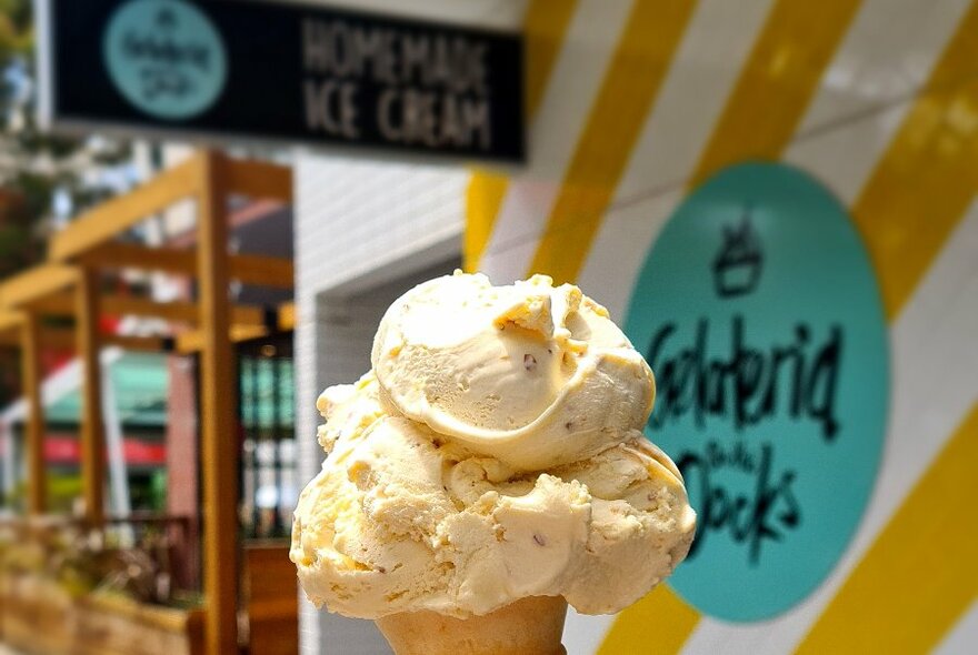 Double scoop ice-cream cone with yellow and white striped Gelateria shopfront in background.