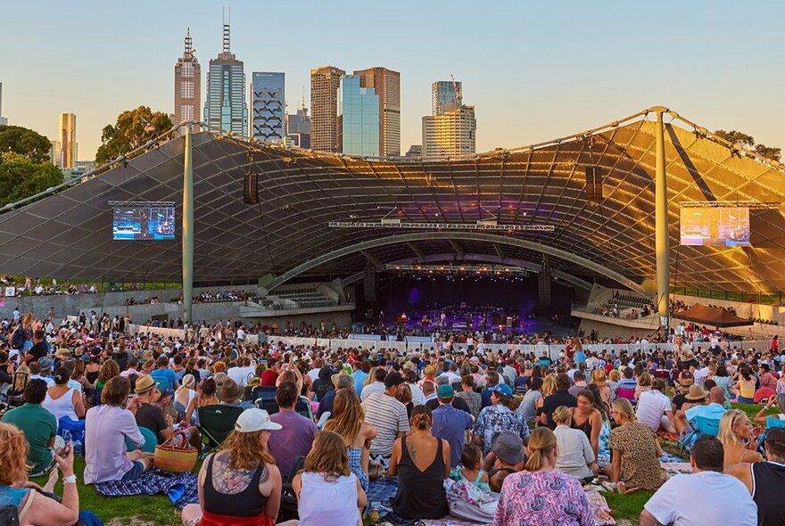 People sitting on the grass in front of a large outdoor stage in the city.