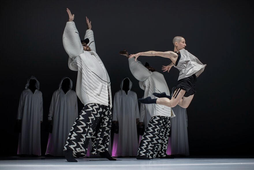 A dynamic modern dance performance on a dark stage, with cloaked figures in the background and three dancers centre stage, one leaping in the air.