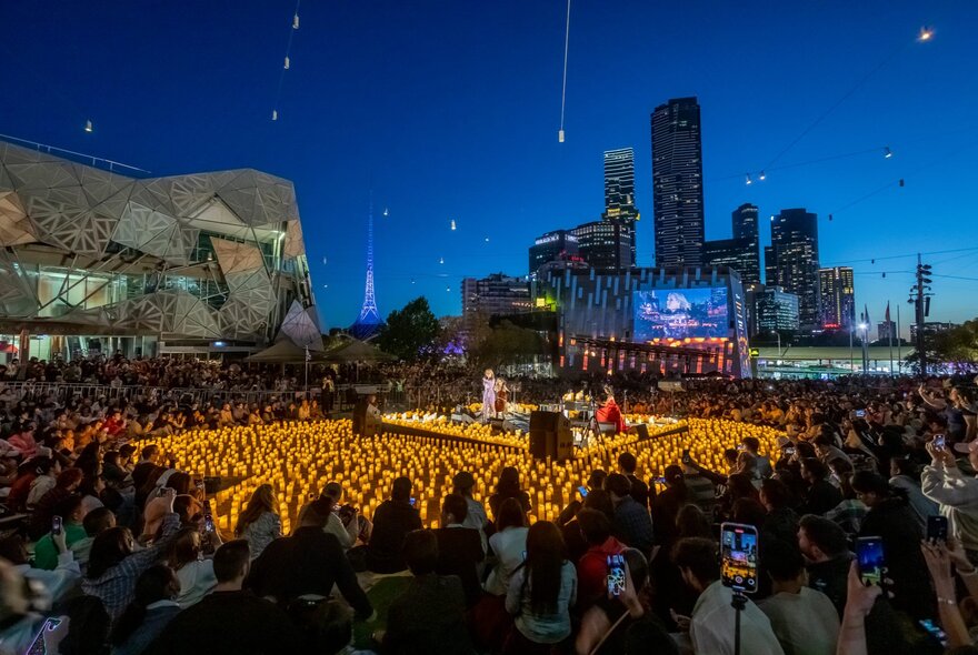 Fed Square at night with twinkling candles and illuminated city buildings in the background.