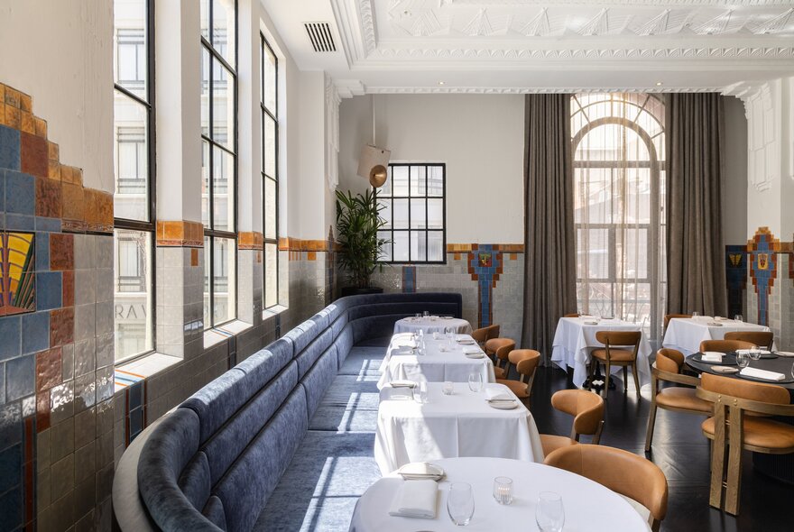 Elegant interior of a restaurant with pale blue banquette seating along one wall, dining tables covered in white linen, high ceilings and large windows, with sunlight streaming into the room.