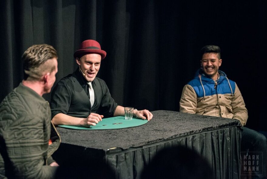 Three men on stage, seated, the middle one performing a magic trick. 