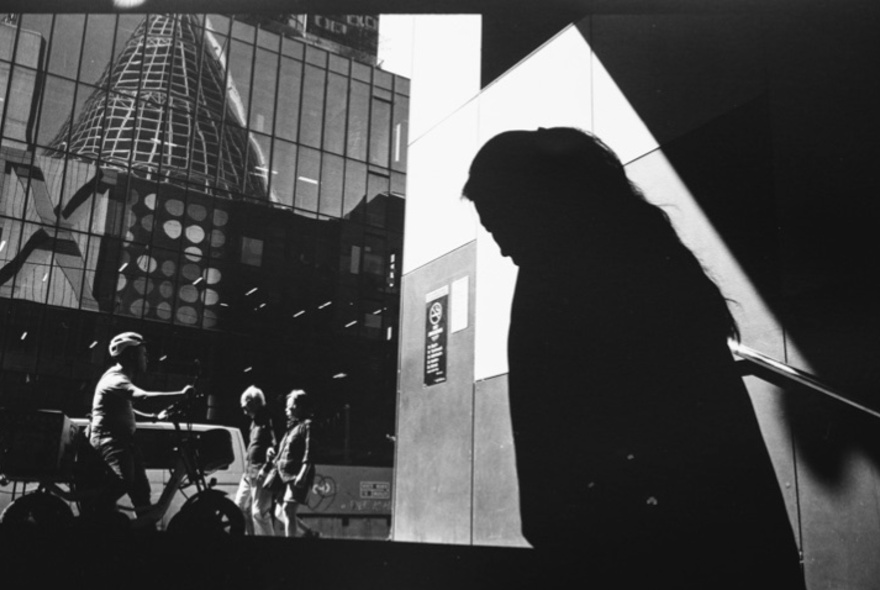 Shadowed figure walking up steps to join people dwarfed by city buildings.