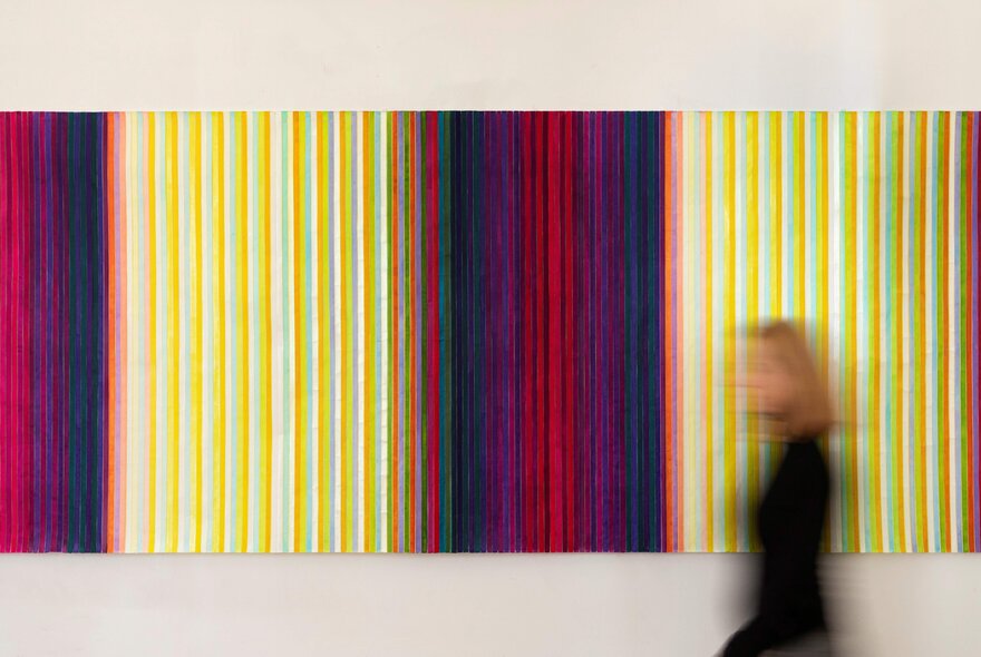 An out-of-focus person walking past a large rectangular artwork hanging on a wall, the artwork comprised on vertical stripes of varying colours.