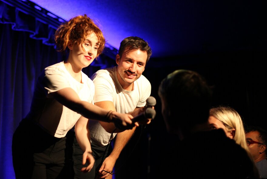 Two comedians on a stage, holding a microphone in front of an audience member they are interacting with.