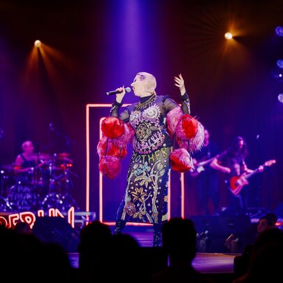 A cabaret perfomer singing on stage before a seated audience; purple lights on her.