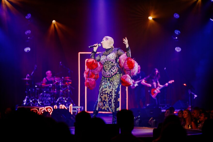 A cabaret perfomer singing on stage before a seated audience; purple lights on her.