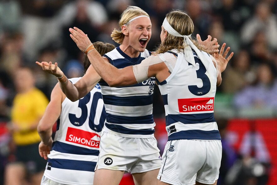 Three Geelong AFL players celebrating on the field after having scored, with blurred fans in the stadium in the background.