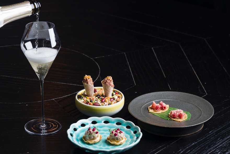 Sparkling wine being poured into a glass, with three dishes of food, arranged on a dark restaurant table.