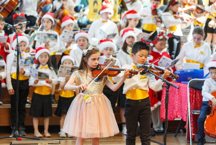 Two young children playing violin with a choir of children singing behind them, some wearing festive red Christmas hats.