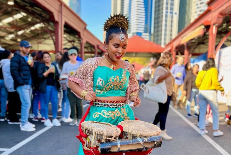 A woman in Sri Lankan traditional dress playing drums at the Queen Victoria Market with shoppers nearby.