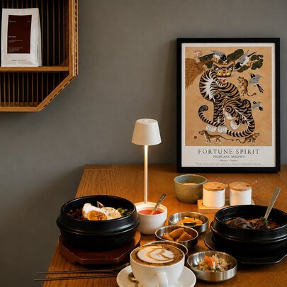 A wooden table in a restaurant with an array of dishes, a cup of coffee, a small table lamp, and an artwork resting against the wall.
