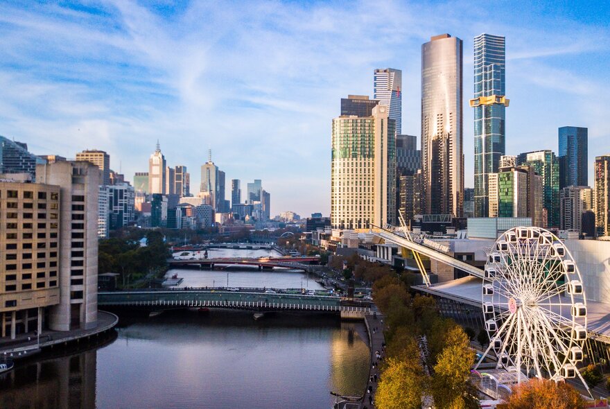 Skyline Melbourne Ferris wheel in the right foreground, the Yarra river with bridges and a skyline of skyscrapers and other large buildings, against a blue sky.
