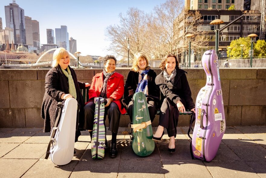 Four female classical musicians holding stringed instrument cases, seated on a bridge against a grey brick wall with city in the background.