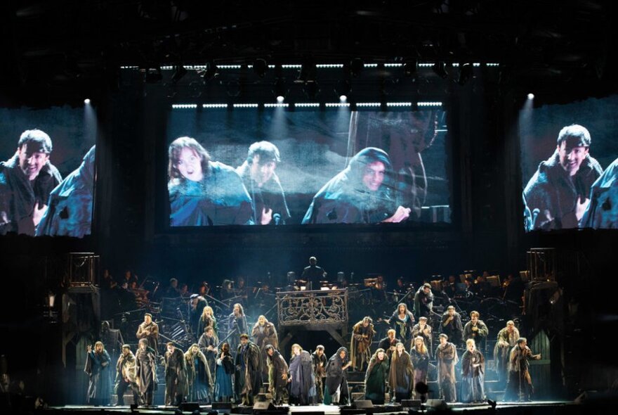 Large ensemble cast performing on stage in the musical Les Misérables The Arena Spectacular, with large screens at the rear of the stage projecting the singers for the audience.
