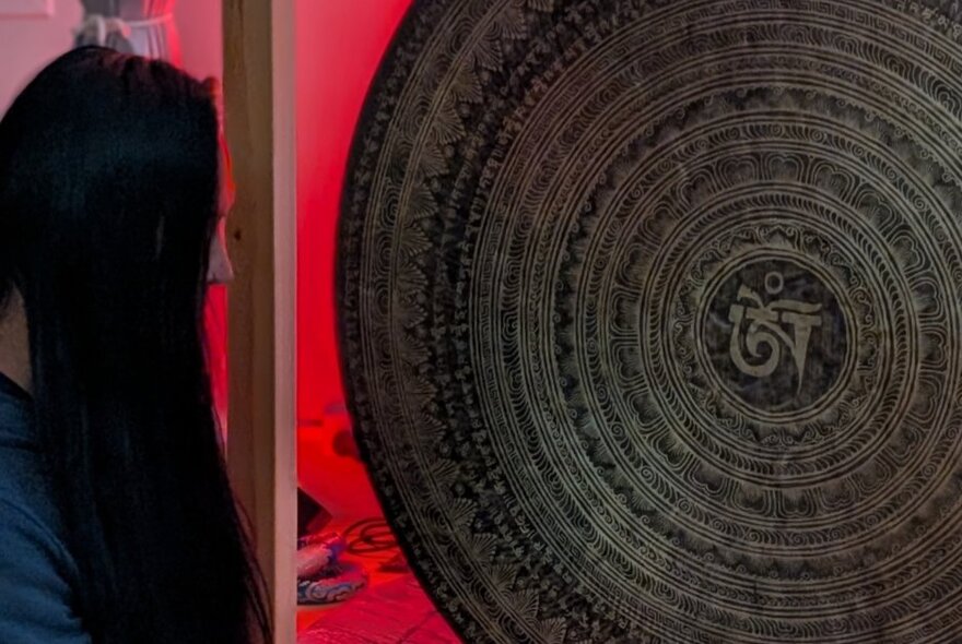 A large, round, dark bronze meditation gong featuring an engraved Om symbol at the centre surrounded by concentric decorative rings, with a figure in profile to the left of the gong.