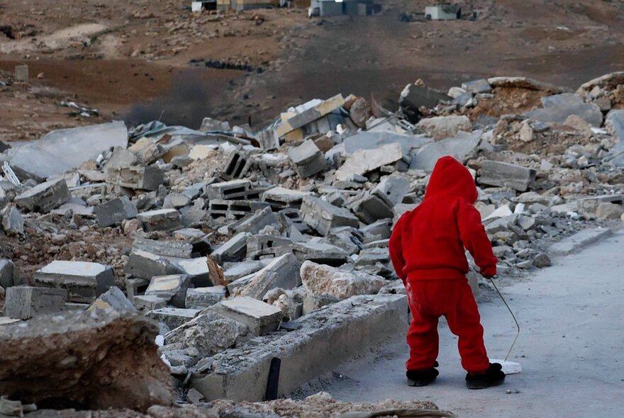 Still from the documentary film No Other Land showing a young child dressed in red playing in the street, surrounded by building and concrete rubble.