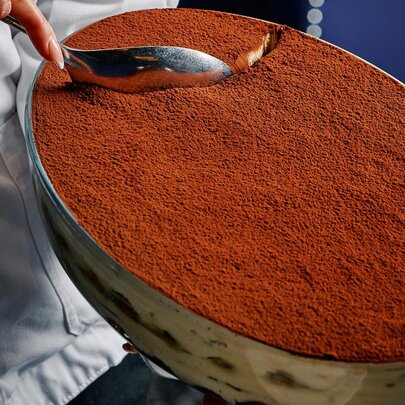 A large oval-shaped bowl of tiramisu, a large silver spoon scooping it.