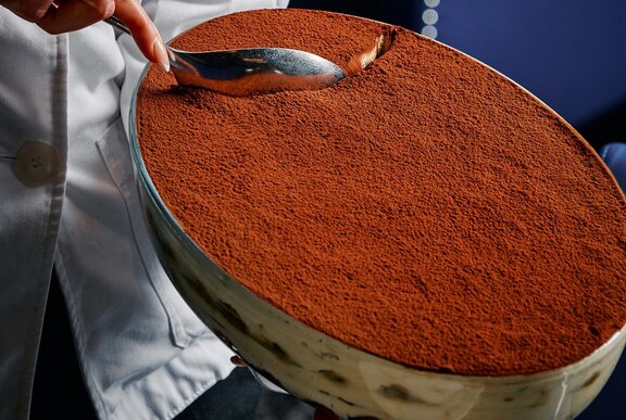 A large oval-shaped bowl of tiramisu, a large silver spoon scooping it.
