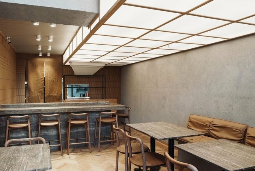 Modern and minimal interior of Waku Waku restaurant showing high stools at a kitchen counter, a small serving area behind this, and dining tables to the side against a side wall. 