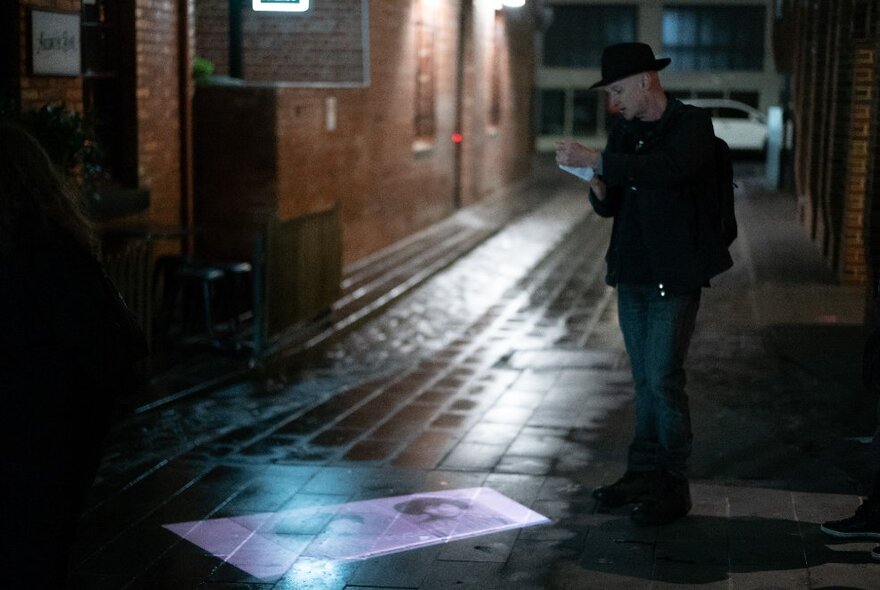 A man wearing a black hat shining a projection  from a hand held projector, onto wet bluestones in a laneway.