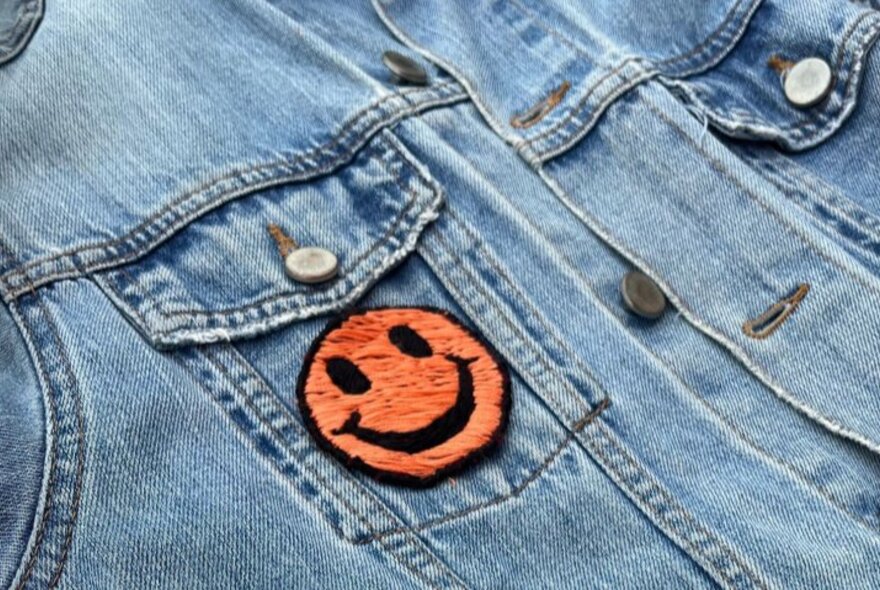 An orange and black smiley face embroidered patch on the pocket of a denim jacket.