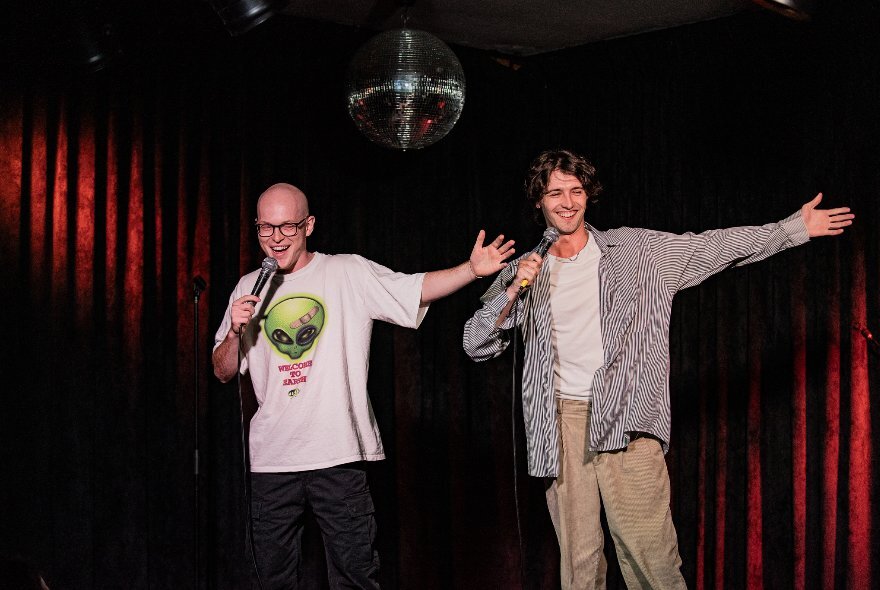 Two comedians on stage, both holding microphones with their right hands and holding out their left arms.