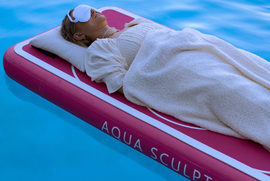 Person lying on a floating bed on a pool, with eye mask and pillow.