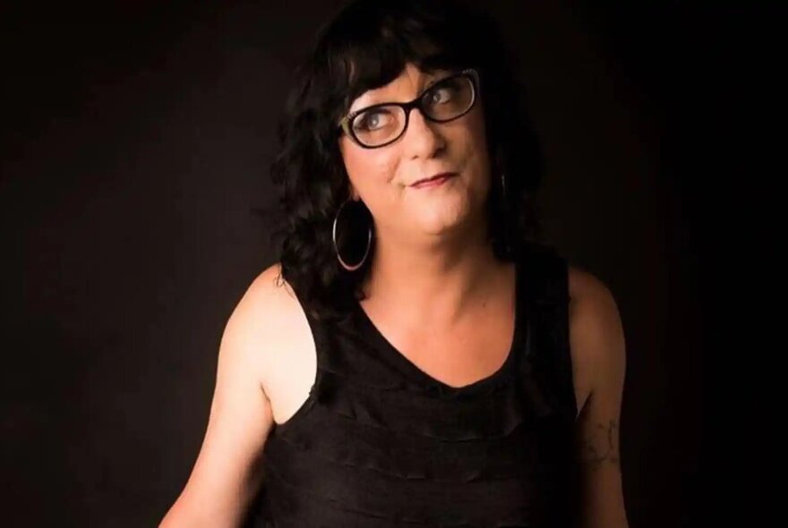 A person wearing a black sleeveless top and glasses, standing in front of a dark background.