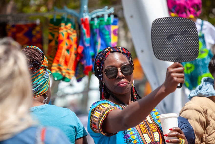 A woman dressed in a blue traditional African dress holding a black perforated fan in front of her.
