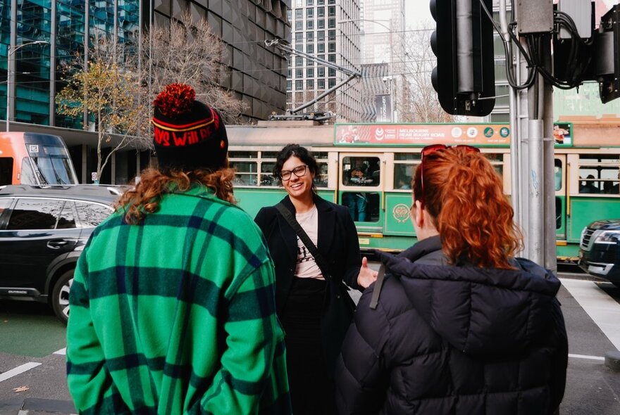 A tour guide talking to two people at a Melbourne city street intersection, a vintage class W tram, cars and tall buildings in the background.