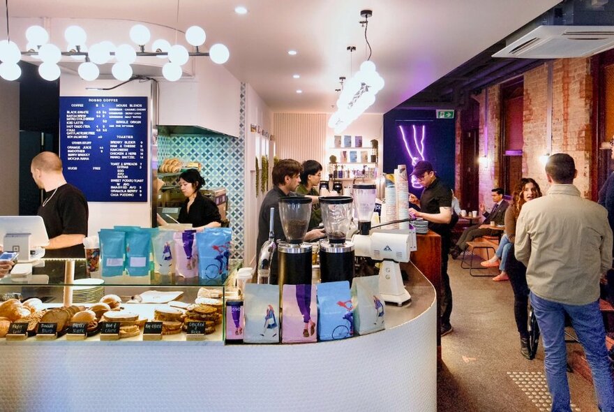 The curved counter of a cafe with baristas working, people waiting and sandwiches and cakes behind a glass screen