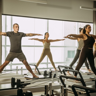 Reformer Pilates class at a KĀYA health club, featuring people standing on adjustable resistance beds in a large airy room.
