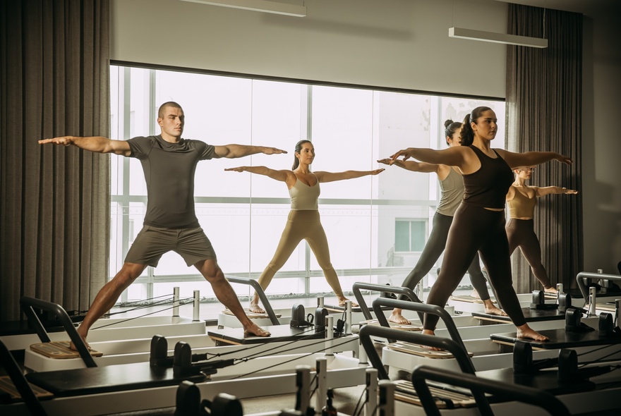 Reformer Pilates class at a KĀYA health club, featuring people standing on adjustable resistance beds in a large airy room.